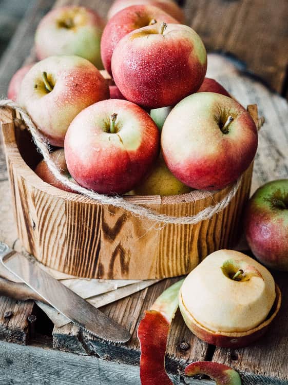 Apfelbaum Seidenhemdchen kaufen - zahlreiche Äpfel in einem Holzkübel auf einem Tisch, mit geschältem Apfel daneben